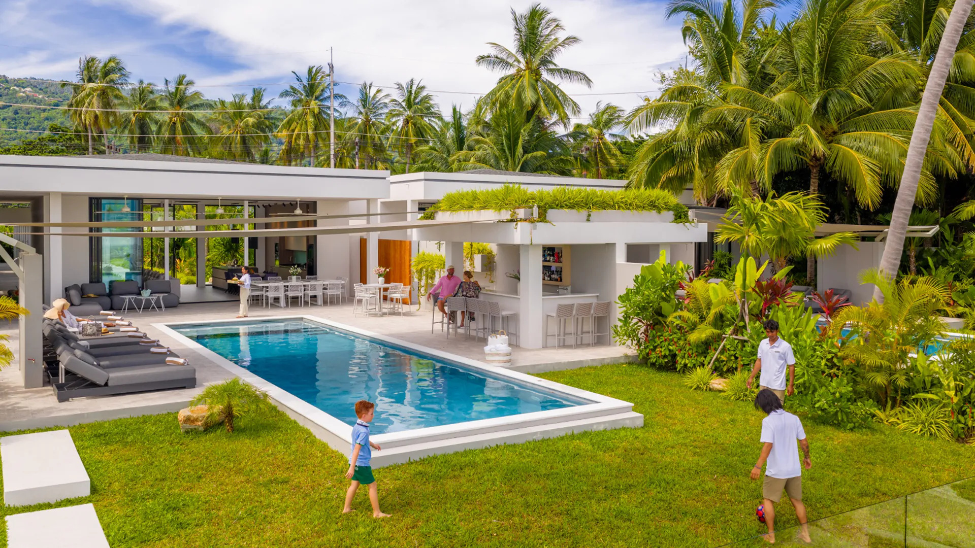 A Private Beach Estate Sanctuary in Koh Samui A family enjoys the tranquil garden area of their luxurious private beachfront villa in Koh Samui, with lush landscaping surrounding them at sunset.