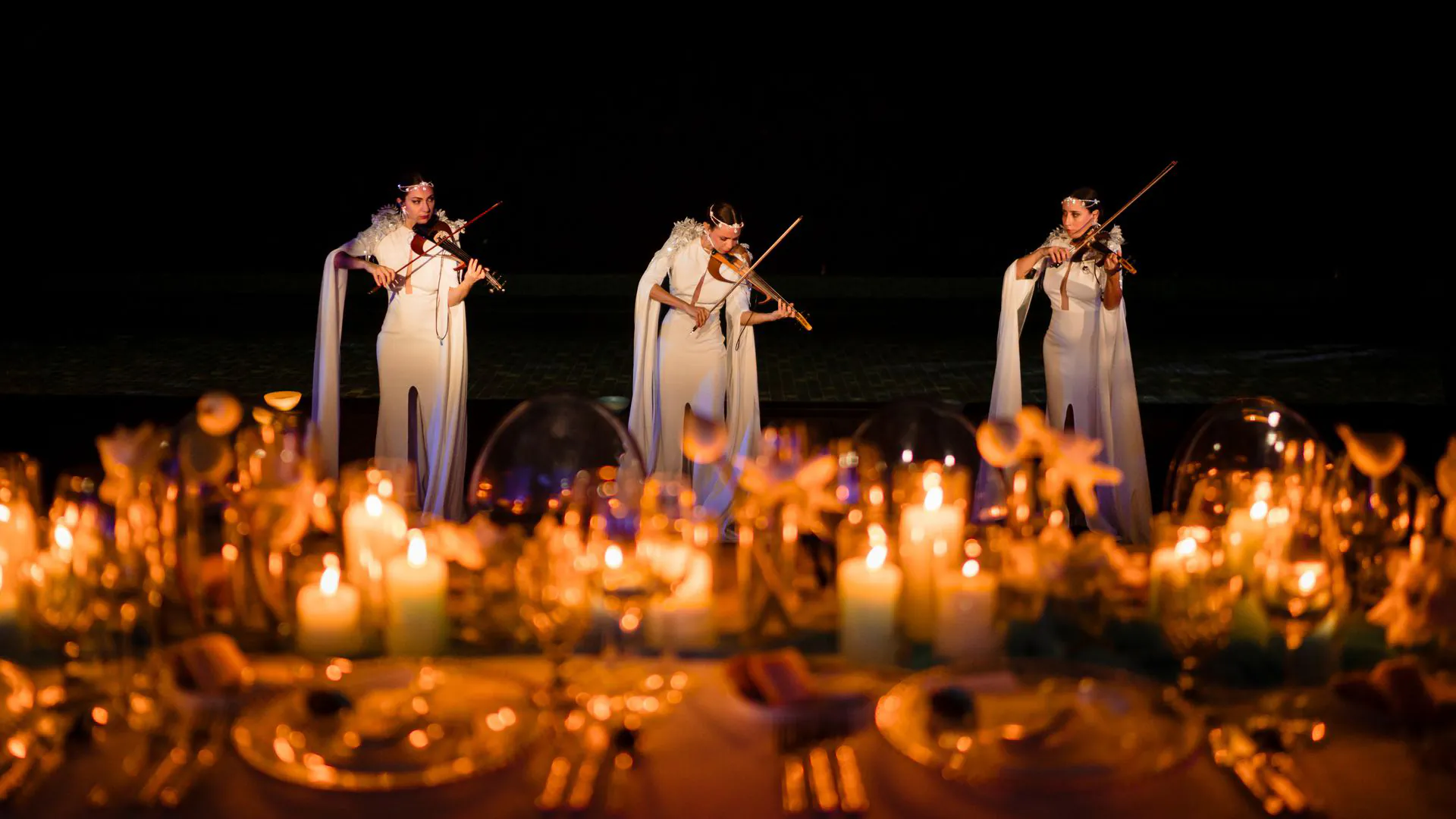 Three violinists in flowing white capes perform at an evening candlelit wedding ceremony at Shankara Estate, Samui, surrounded by hundreds of glowing candles creating an intimate atmosphere for a large-scale luxury wedding.