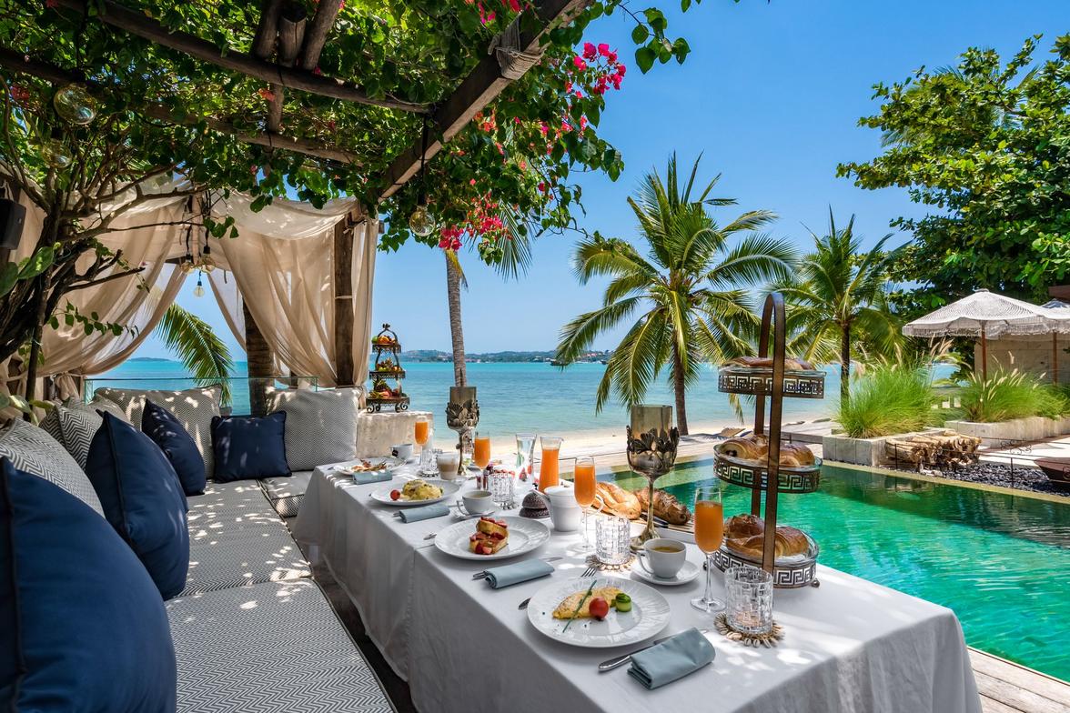 A stunning luxury modern Koh Samui villa features an outdoor dining area under a blooming pergola, with breakfast set overlooking a cinematic architecture of the private pool and the sparkling turquoise ocean.