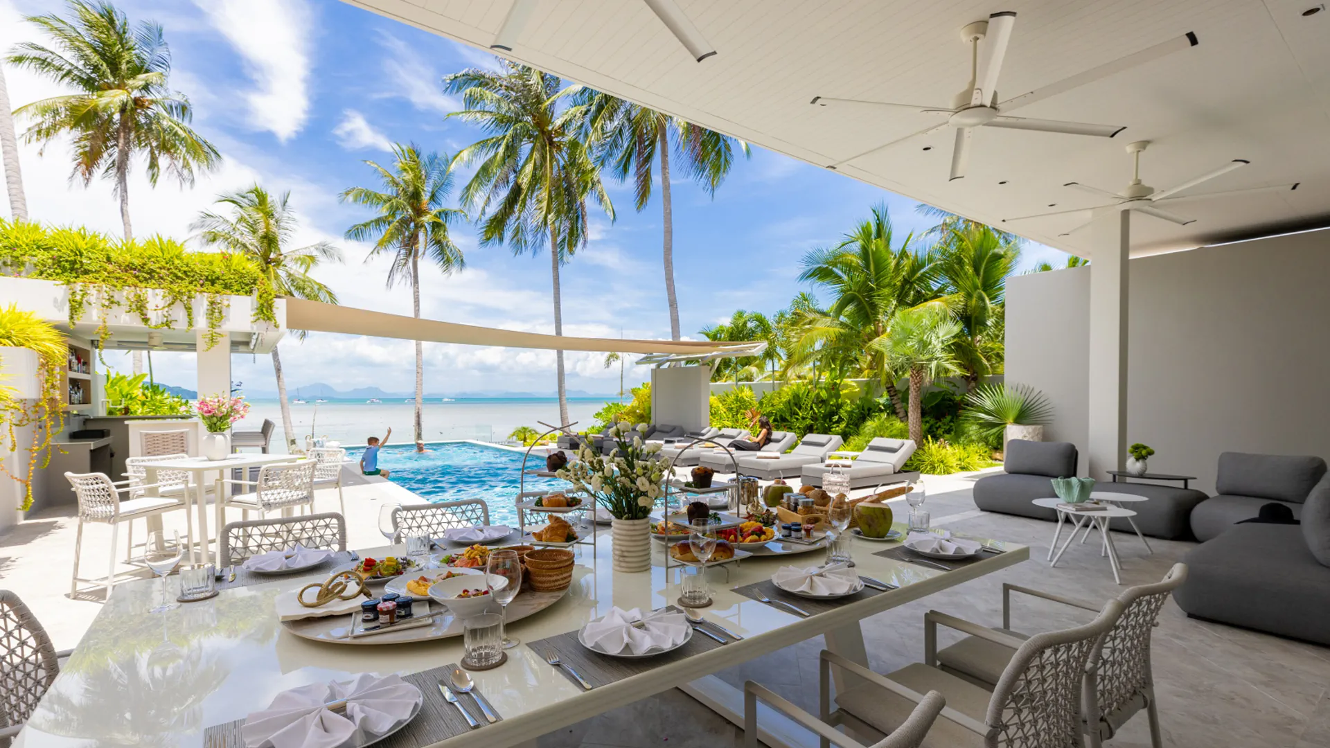 Synchronized Chef Program Beachfront Dining at Emotion Beach Villas, Koh Samui Luxury outdoor dining table set for breakfast at Emotion Beach Villas with ocean views, infinity pool, palm trees and white modern furniture on covered terrace.