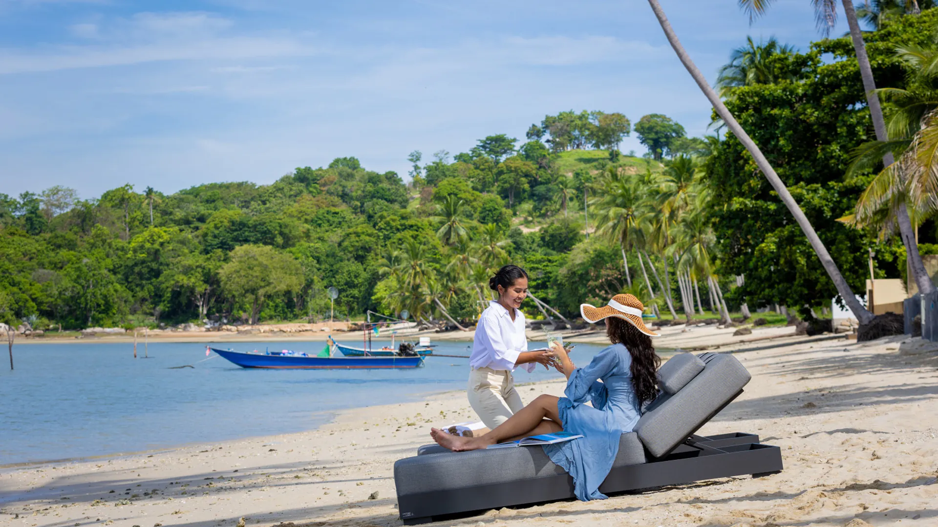 Beachfront Relaxation at Emotion Beach Villas - Personalized Service Two guests relaxing on beach loungers at Emotion Beach Villas estate in Hua Hin, with traditional longtail boat and palm-lined shore in background.