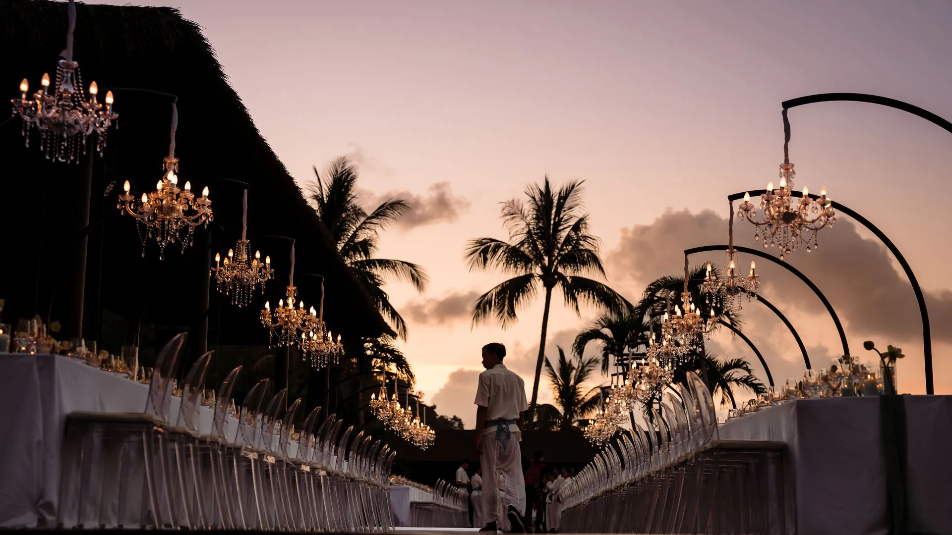 Elegant outdoor wedding setup at sunset with crystal chandeliers suspended over white-draped tables, palm trees silhouetted against pink sky at tropical beachfront venue.