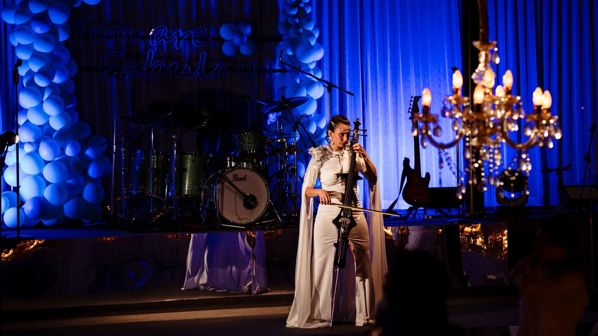 Professional cellist performing at an elegant wedding reception in Thailand with blue ambient lighting, balloon decorations, and crystal chandelier creating a luxury atmosphere.