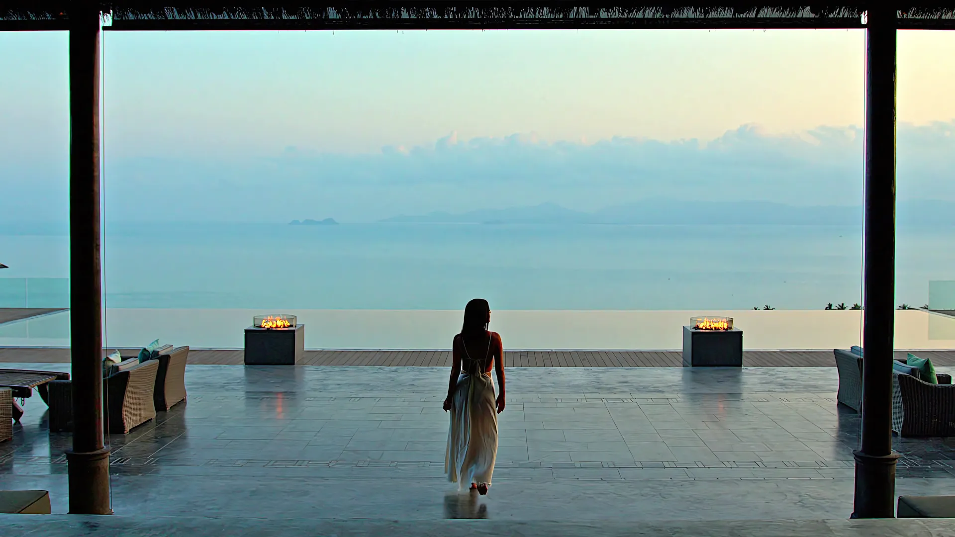 Orchestrating an Unforgettable Guest Arrival Experience Guest in flowing dress walking through grand open-air pavilion towards infinity pool with fire features at sunset, showcasing dramatic arrival experience at Private Estate Shankara, Maenam, Koh Samui.