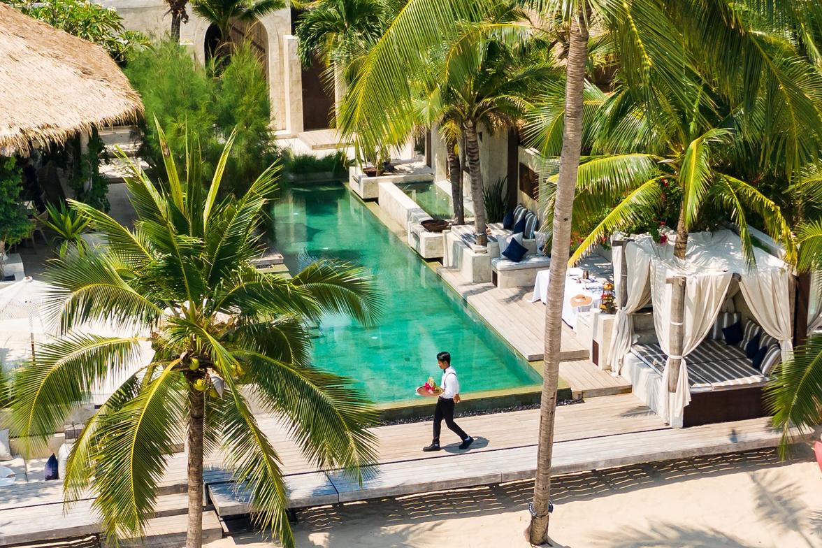 The Pinnacle of Private Luxury in Koh Samui A private luxury resort on a beach in Koh Samui with a long, emerald swimming pool, surrounded by palm trees and cabanas, as a staff member walks by.