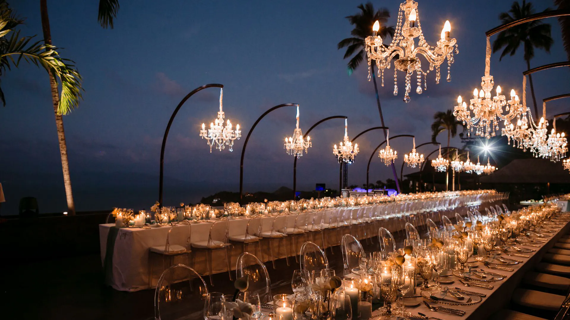Elegant beachfront wedding reception at dusk with long banquet table, crystal chandeliers suspended from arched stands, candlelight, and palm trees silhouetted against twilight sky.
