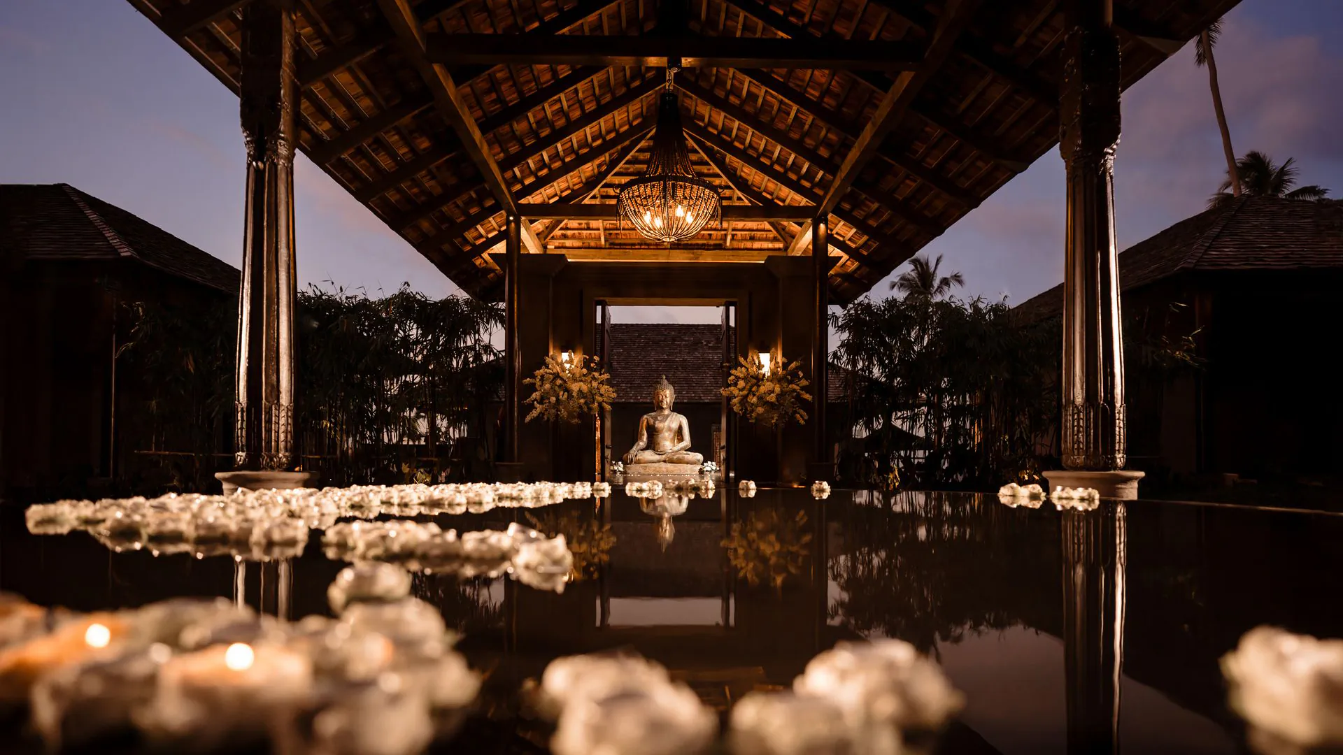Shankara Estate wedding pavilion at dusk with Buddha statue, floating candles on reflecting pool, and traditional Thai architecture with ornate chandelier.