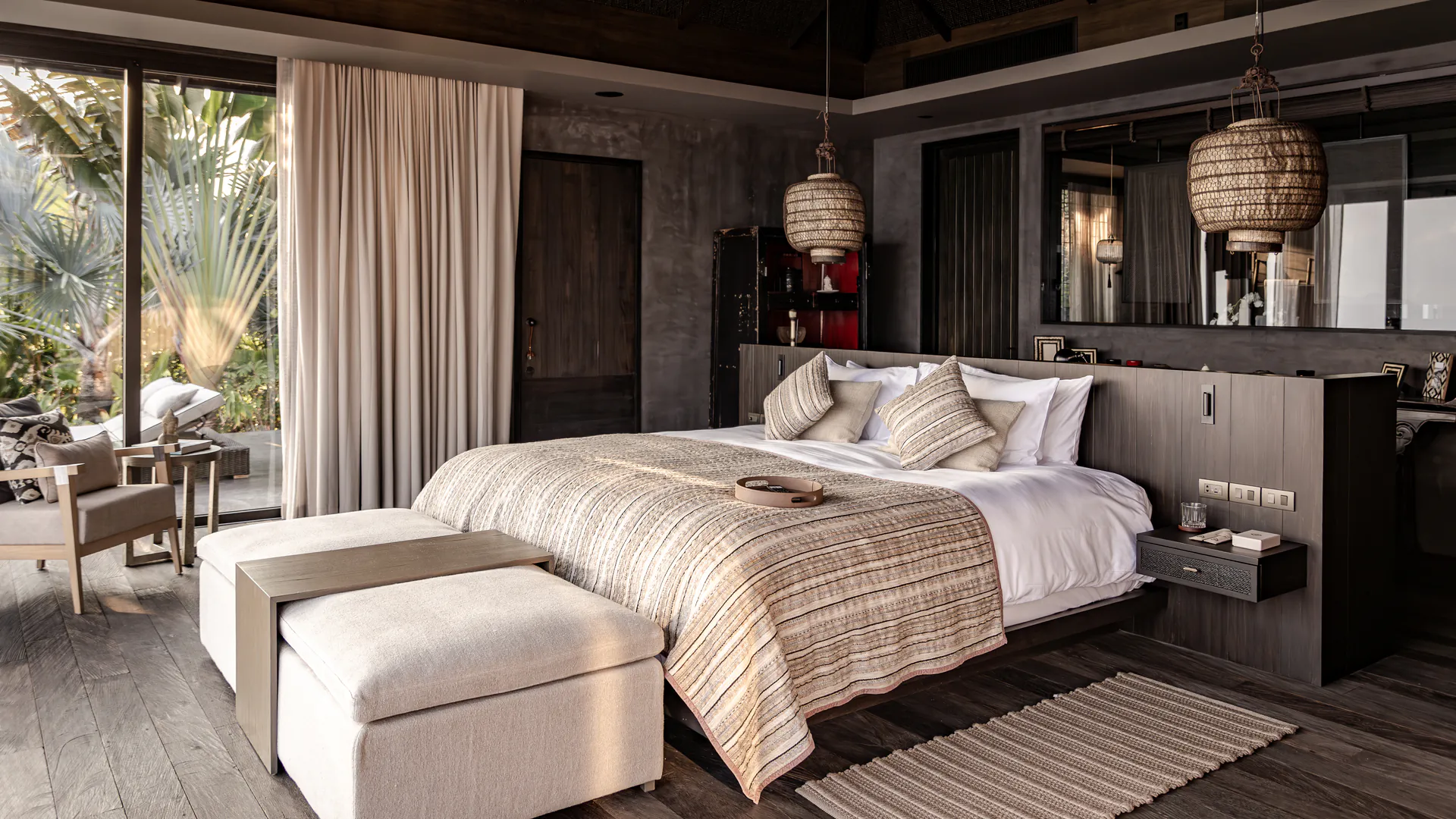 Luxurious master bedroom suite at Shankara Estate featuring dark wood floors, handwoven pendant lights, neutral textured bedding, and floor-to-ceiling windows opening to tropical garden views.