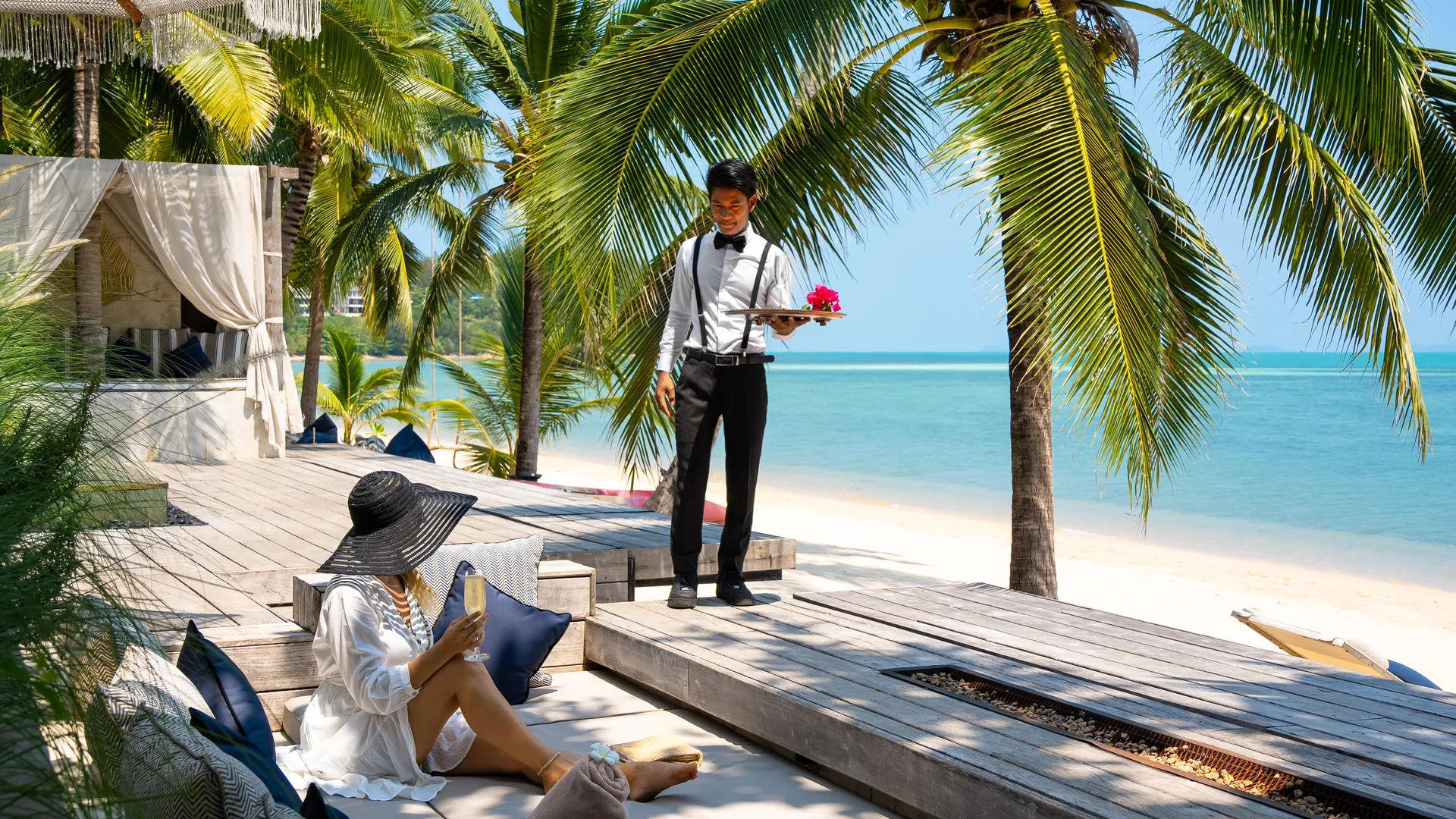 The Invisible Standard of Luxury Villa Service Butler in formal attire serving a guest relaxing with a book on a wooden deck overlooking turquoise waters, surrounded by palm trees at Villa Odyssey, Bangrak, demonstrating discreet luxury service in tropical Koh Samui